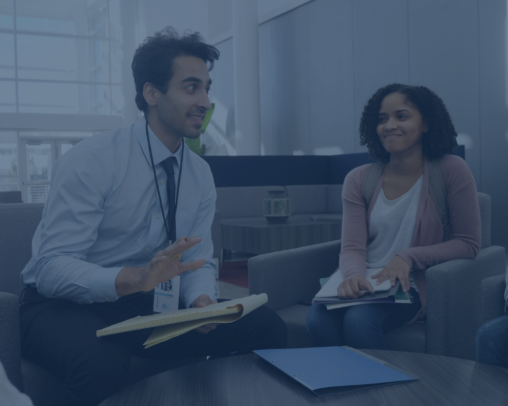 Image of teachers talking. Male teacher leader is animatedly speaking. Black teacher is smiling and listening as they discuss how to submit scores on curriculum implementation.